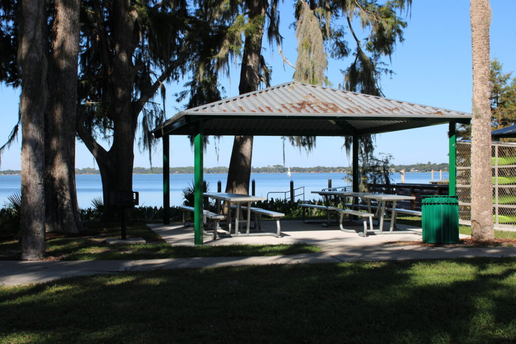 A shaded picnic pavilion with tables near the Palm Island Boardwalk, overlooking a sailboat on Lake Dora.