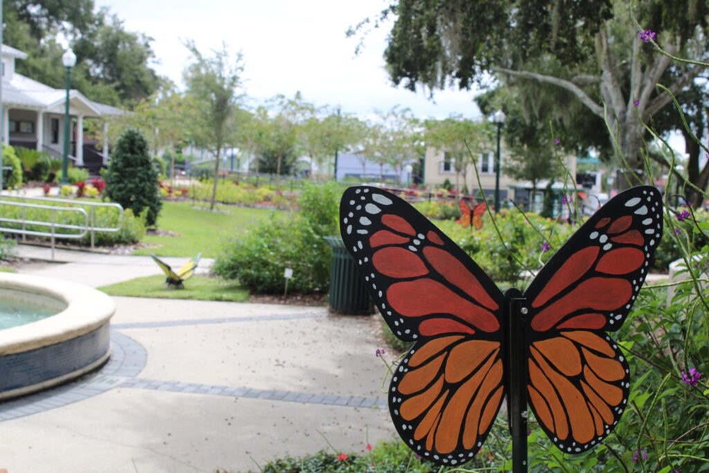 A brightly colored, large monarch butterfly statue painted orange and black, seen up close near the paved fountain area with the Community Building visible in the background.
