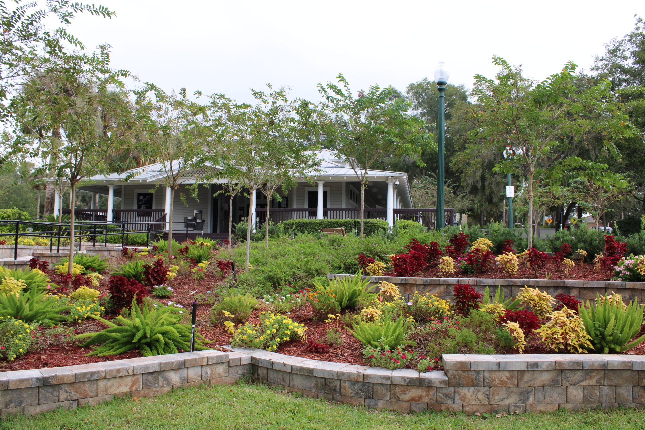 Wide view of the historic white Donnelly Park Community Building, set back on a landscaped hill with young trees and bright red and yellow foliage, serving as the central hub of the downtown park.