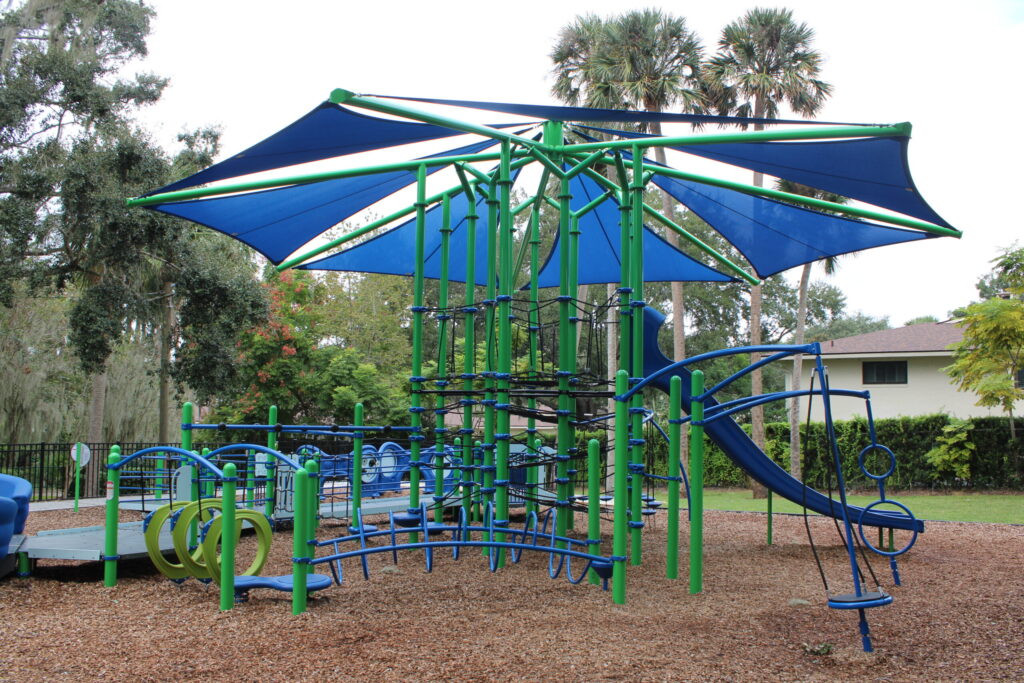 A blue and green shaded playground structure with slides and climbing nets for toddlers at Gilbert Park.
