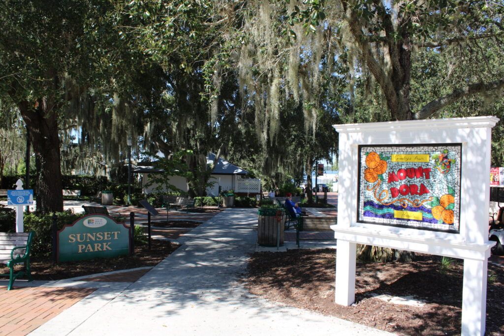 Downtown Mount Dora's Sunset Park main plaza featuring the paved square, central flag pole, decorative planters with bushes, and the historic white facade of the Mount Dora Chamber of Commerce and Visitor Center building under a bright blue sky.