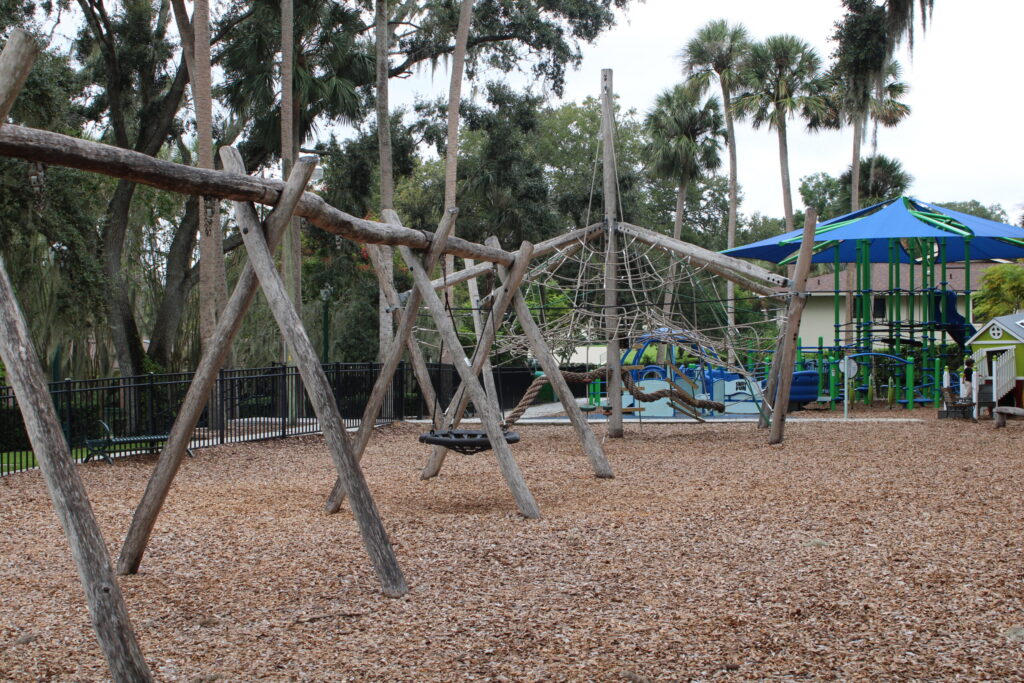 Swings and climbing nets made of natural wood at the fenced Gilbert Park playground in Mount Dora, FL.