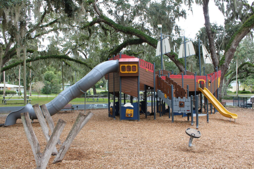 Wide view of the pirate ship playground structure under large, mossy oak trees at Gilbert Park, Mount Dora, FL.