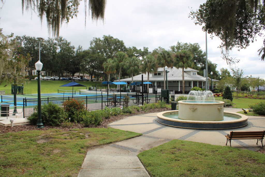Close-up view of the tiered water fountain with jets, surrounded by an octagonal paved walkway, a wooden bench, and large shade trees covered in Spanish Moss.
