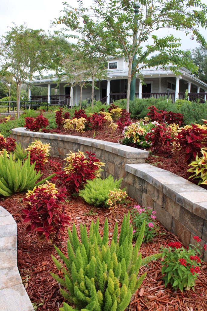 A close-up view of the historic white Community Building porch and columns, framed by a stone retaining wall and lush garden beds filled with vibrant red and yellow coleus plants and spiky green ferns.