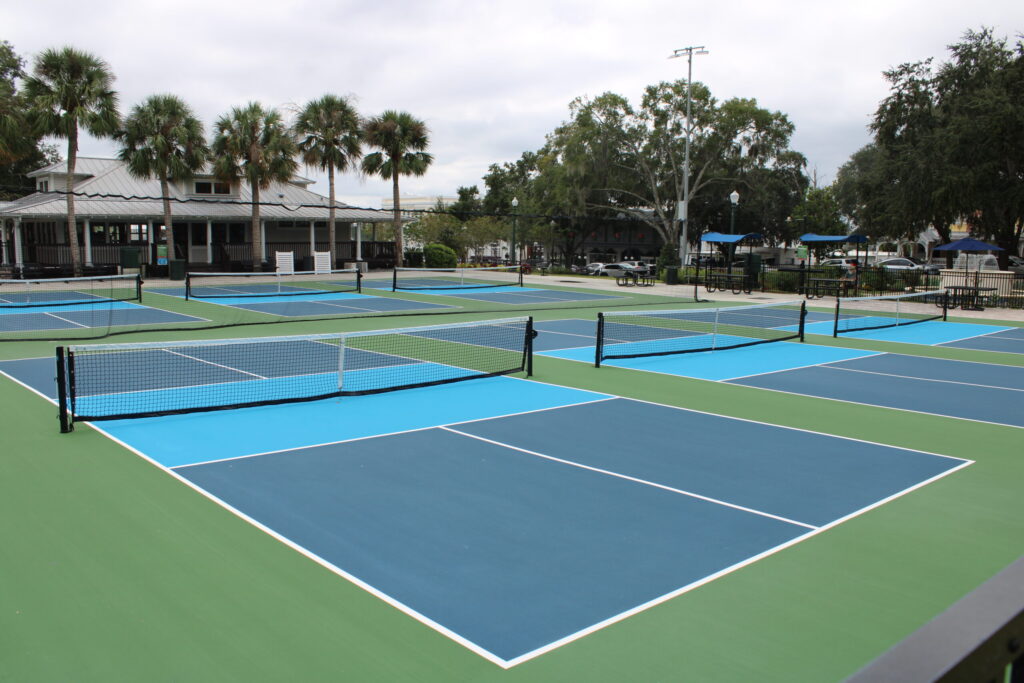 A wide, overhead view of the tiered fountain in Donnelly Park, with a path leading past lamp posts toward the new blue and green pickleball courts in the background, all framed by Spanish Moss on the oak trees.