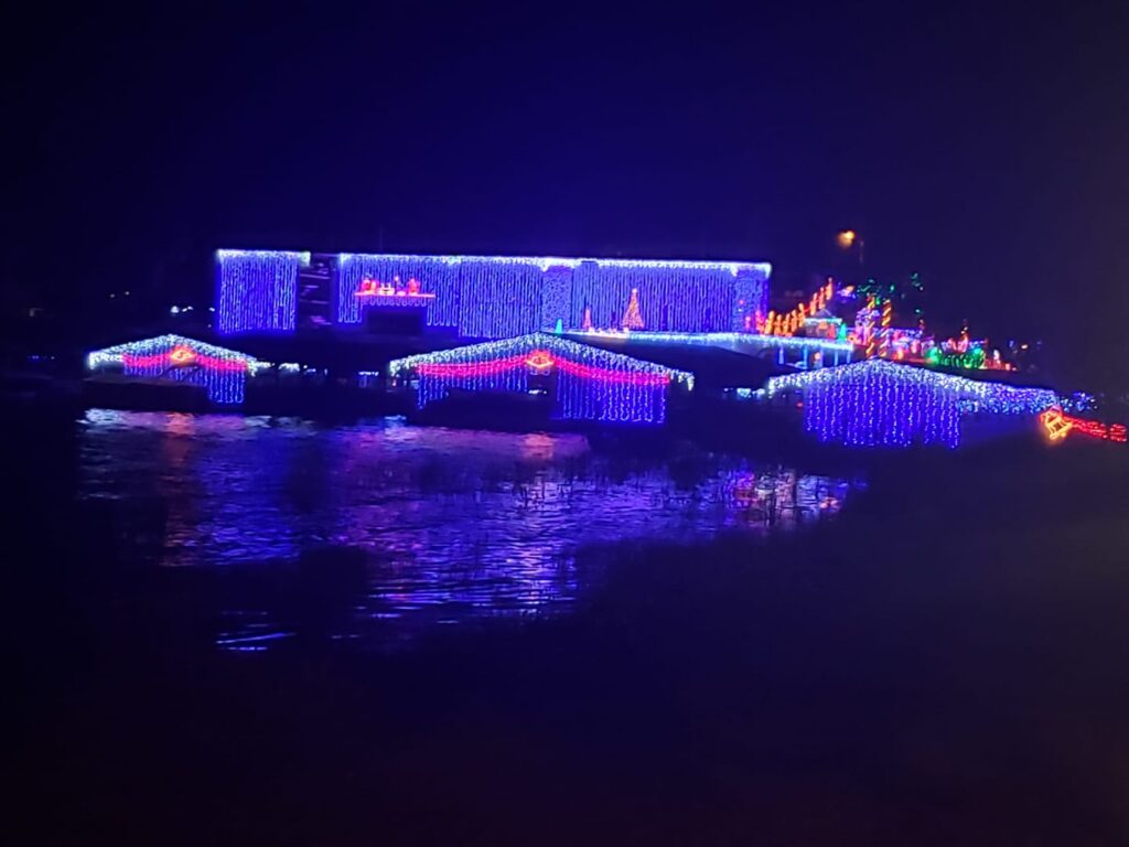 Close-up view of the Mount Dora Marina on Lake Dora at night, where dozens of boat slips are decorated with festive Christmas lights and reflections shimmer brightly on the dark water. The colorful display is part of the annual holiday celebration.
